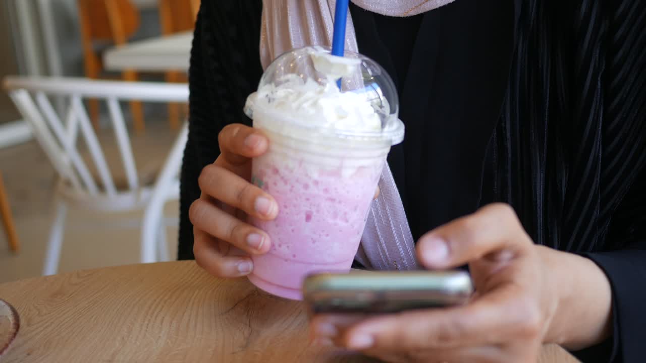 Person holding a pink drink and using a smartphone in a cafe