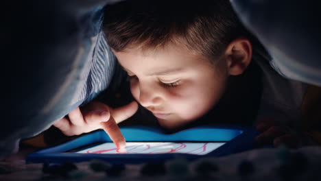 niño feliz usando una tableta digital bajo una manta disfrutando de aprender en la tecnología de pantalla táctil jugando juegos divirtiéndose antes de acostarse