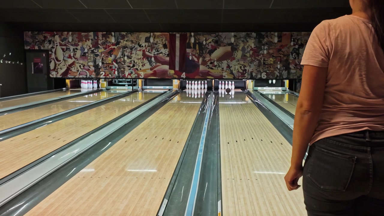 Osoba v růžové košili hraje bowling na vnitřní dráze s postavenými kuželkami a zářivým nástěnným uměním v pozadí v bowlingové herně v Jelgavě, Lotyšsko