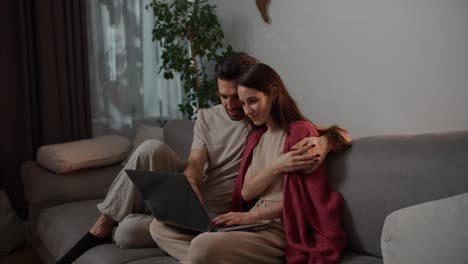 vista lateral de un hombre moreno feliz con paja junto con su joven esposa morena en un suéter rojo sentado en un sofá gris y viendo una película interesante en una computadora portátil gris en un apartamento moderno