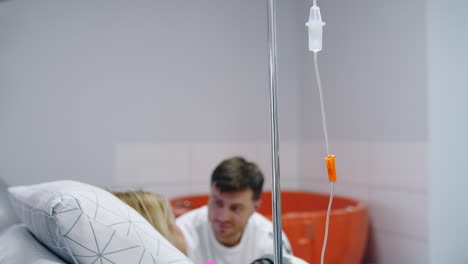 drop counter near the bed of caucasian woman. man talks to his pregnant wife in hospital ward at backdrop in blur.
