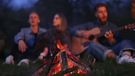 Cozy bonfire burning at nighttime. Friendly campers singing song along with the guitar at the backdrop in blur.
