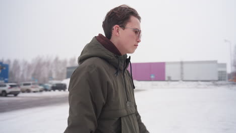 Boy wearing winter jacket with hoodie walks across snowy parking lot toward red car during light snowfall with parked cars around and distant trees lining urban area on cold winter day