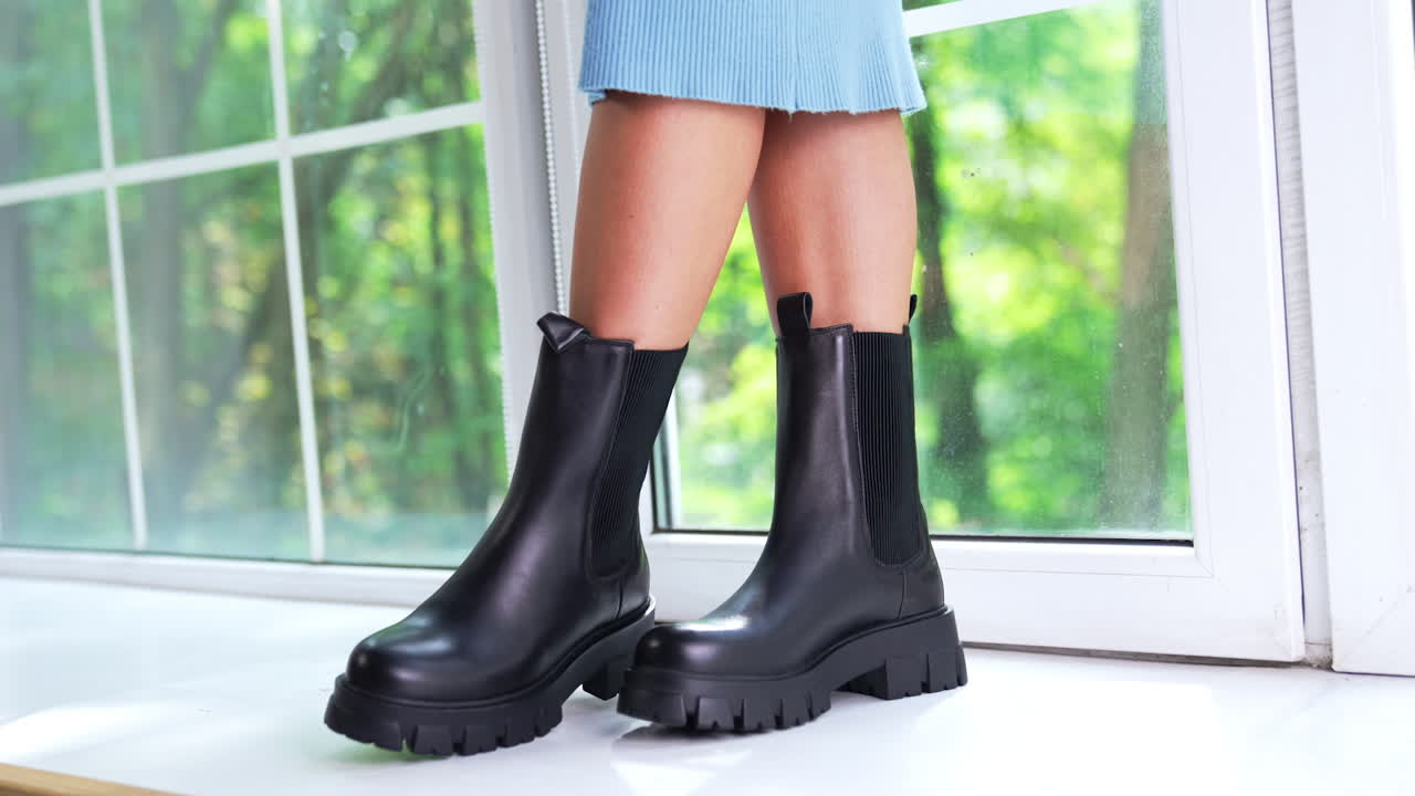 Woman in blue dress wearing black boots. Female model standing on a window-sill demonstrating stylish fashionable leather footwear. Close up.