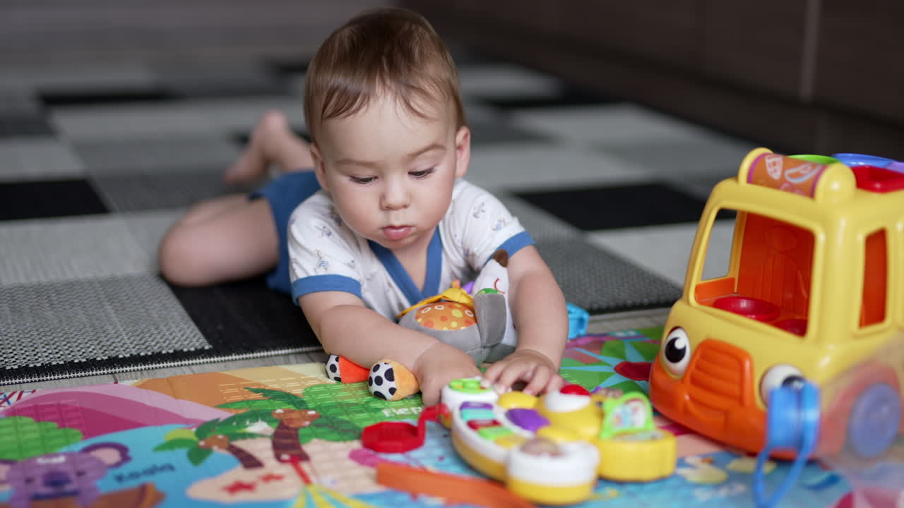 Cute little baby in t-shirt and shorts lies on a carpet on his belly. Lovely toddler playing with toys in front of him. Close up.