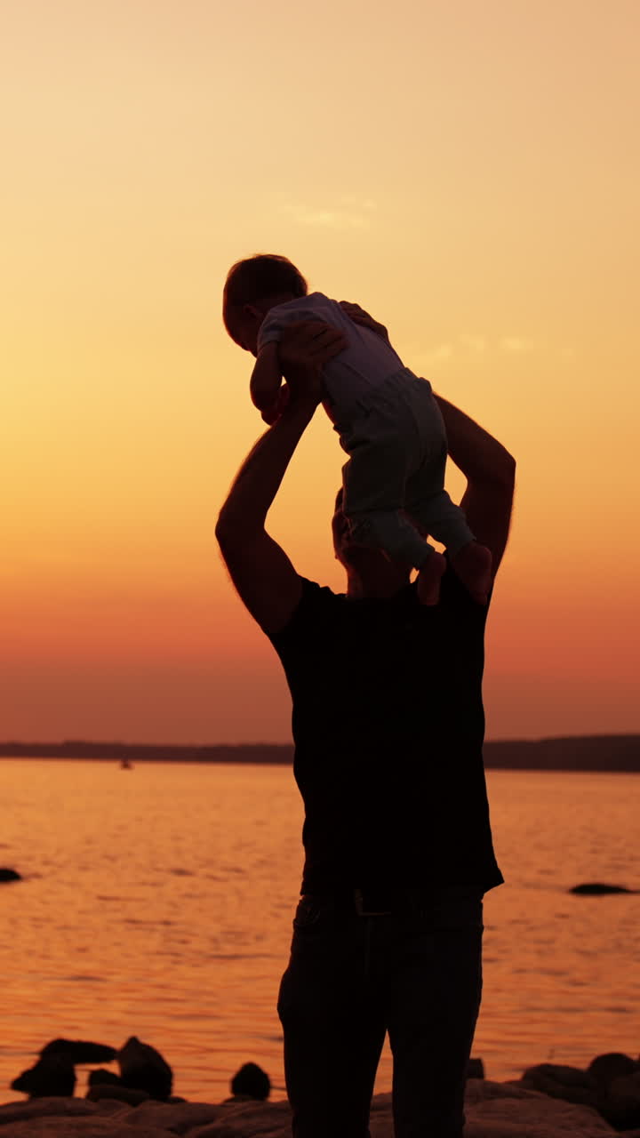 Happy laughing baby boy in the hands of father. Adorable child playing with dad outdoors near the lake at sunset. Vertical video
