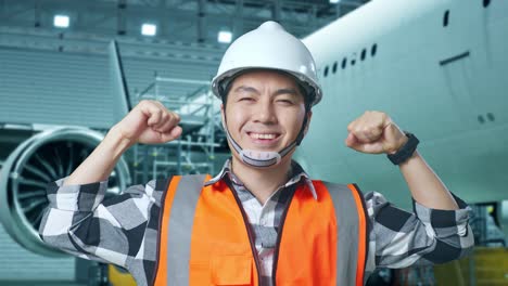 close up of asian male engineer with safety helmet standing with aircraft in the hangar. flexing his bicep and smiling to camera while aircraft maintenance