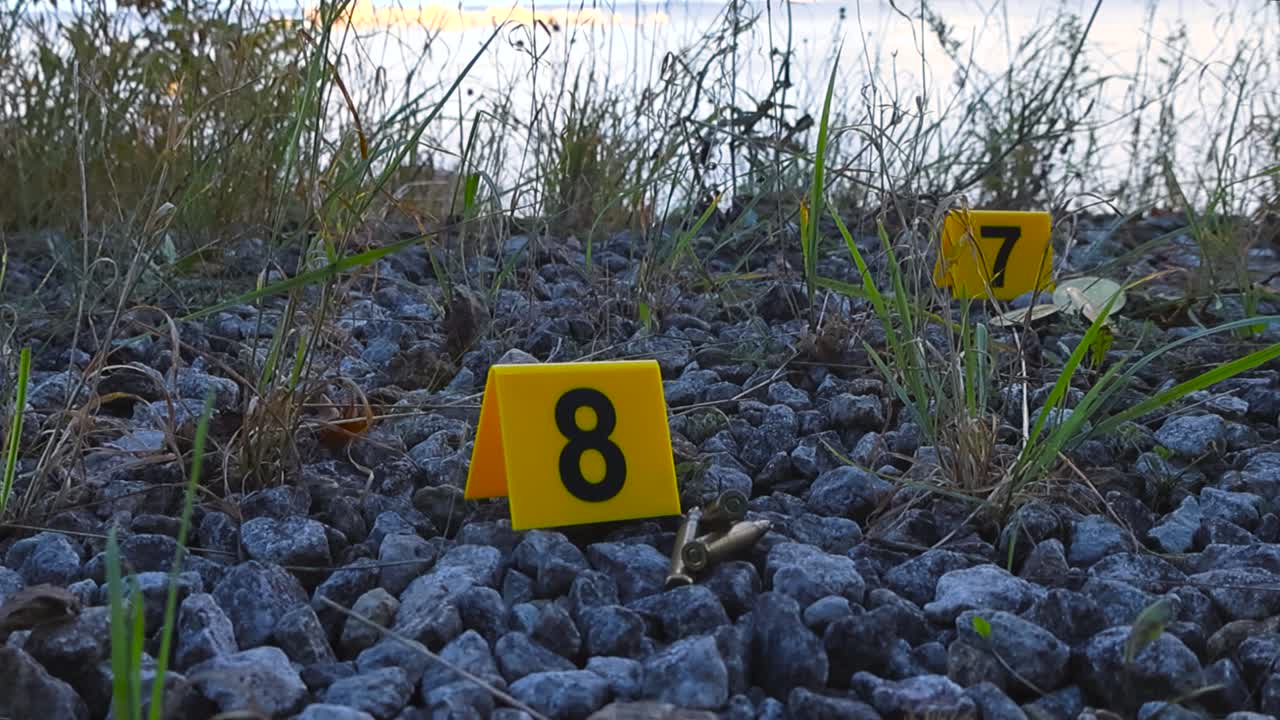 Visualizzazione dal basso di marcatori gialli della polizia scientifica accanto a prove come proiettili, munizioni o munizioni e moneta bitcoin su ghiaia e terreno roccioso in riva al mare