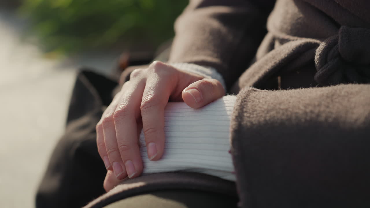 Hands clasped on lap in sunlight, person sits quietly by grassy path with tote bag at side, brown coat and white sleeve framing gentle hand gesture, scene evokes contemplation, mindfulness and calm