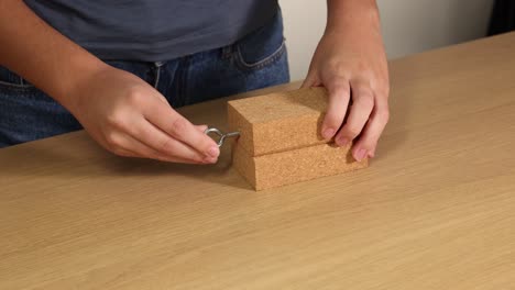 A person inserts a metal hook into a wooden block on a table using hands in a well-lit room