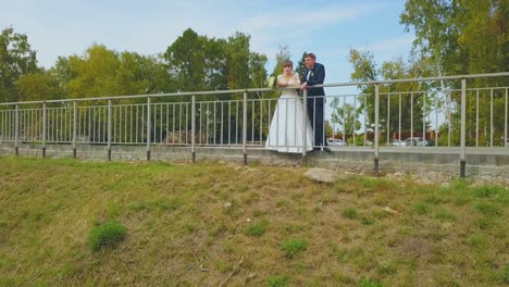 girl-in-wedding-dress-and-groom-lean-on-park-fence-at-lawn
