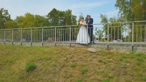 girl in wedding dress and groom lean on park fence at lawn