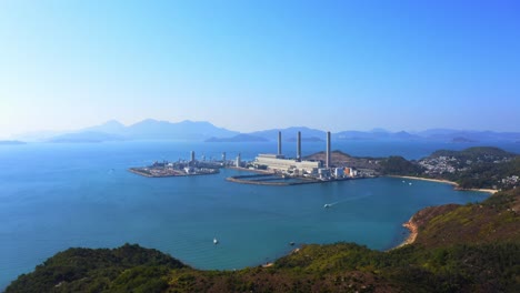 Drone-shot,-traveling-backward,-of-a-big-coastal-coal-fired-power-plant-on-an-island-next-to-a-village-with-a-beach-during-a-beautiful-sunny-day