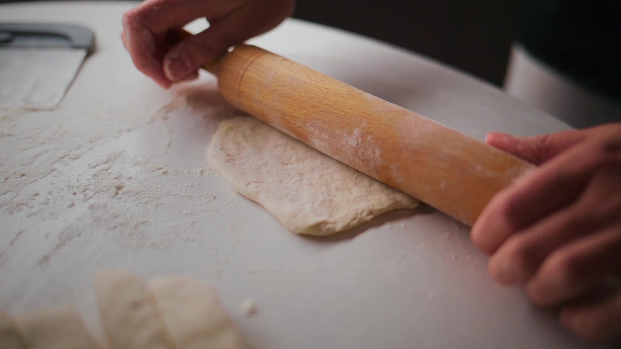 Person rolls dough with wooden roller on floured surface. Hands guide roller gently, ensuring even thickness. Dough stretches slightly under pressure, showing elasticity. Scene suggests home baking