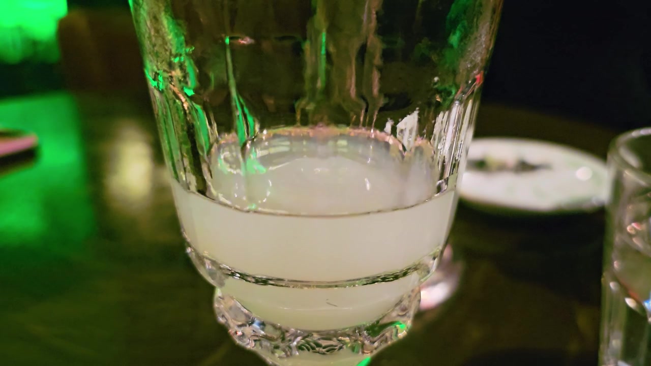 Primer plano macro de absenta louche arremolinándose en un vaso, mostrando una bebida pálida lechosa, textura de cristal tallado, reflejos verdes de la barra, enfoque superficial y un ambiente íntimo de coctelería de noche