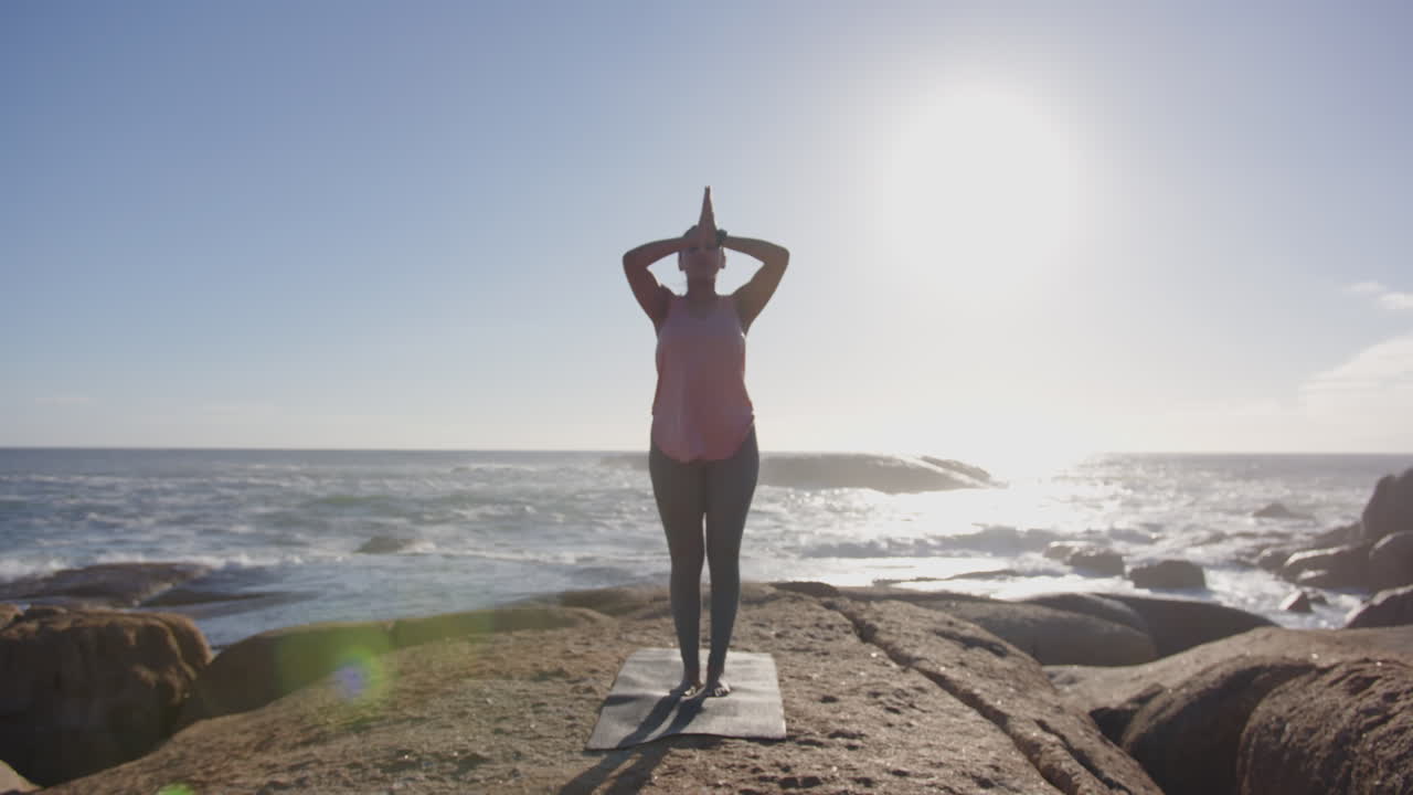 Practicing yoga on mat, mature asian woman meditating by ocean at sunrise