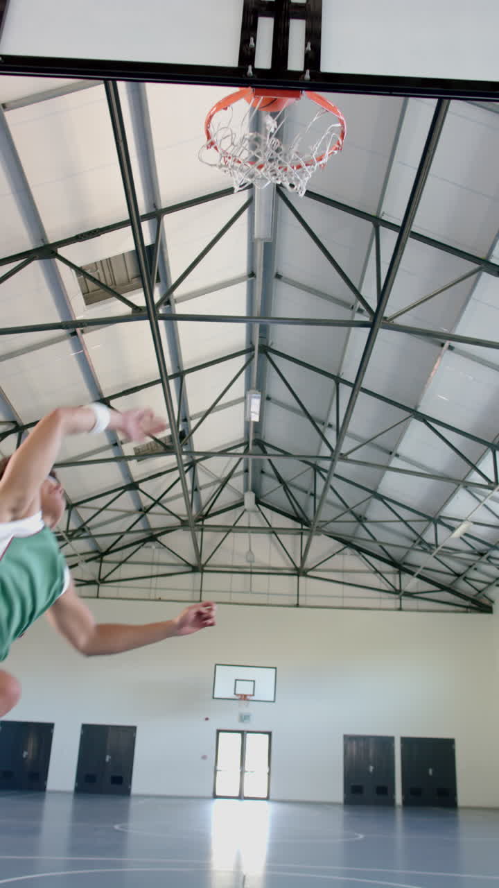 Vertical video: Playing basketball, male athlete dunking ball in school indoor court