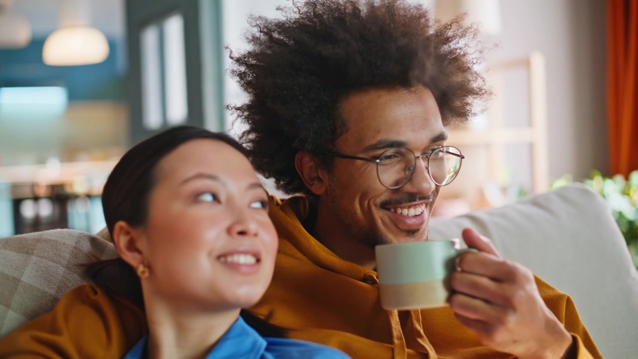 Couple relaxing sofa drinking coffee. Closeup multiracial pair watching TV home.