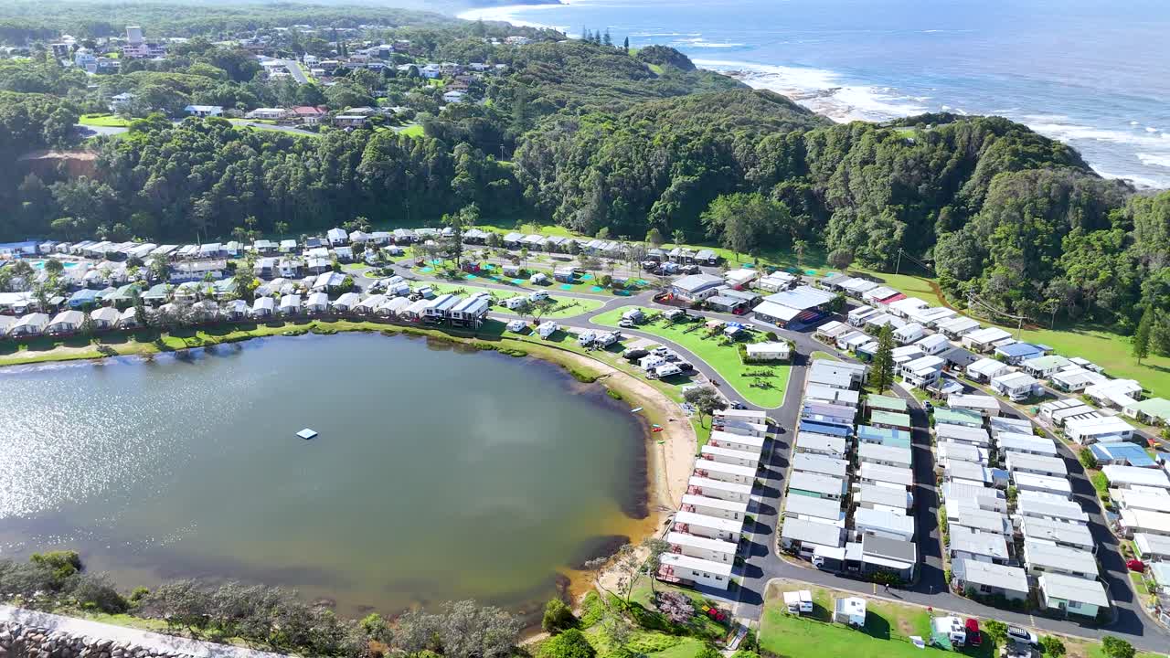 Aerial drone footage over riverside holiday park with cabins, lagoon, forest, and coastline in daylight