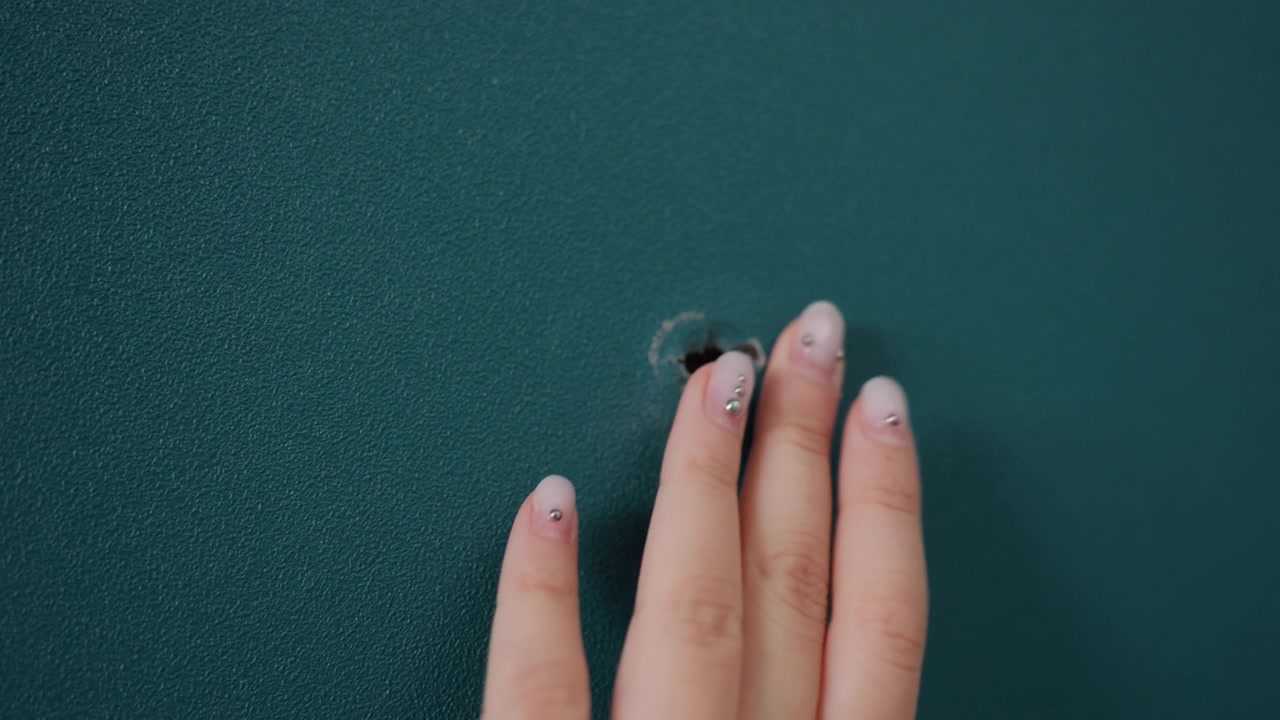 Closeup hand inspecting small hole in teal apartment drywall after moving delicate fingers probe chipped paint and torn drywall paper interior scene shows textured paint, damage near nail point, mood