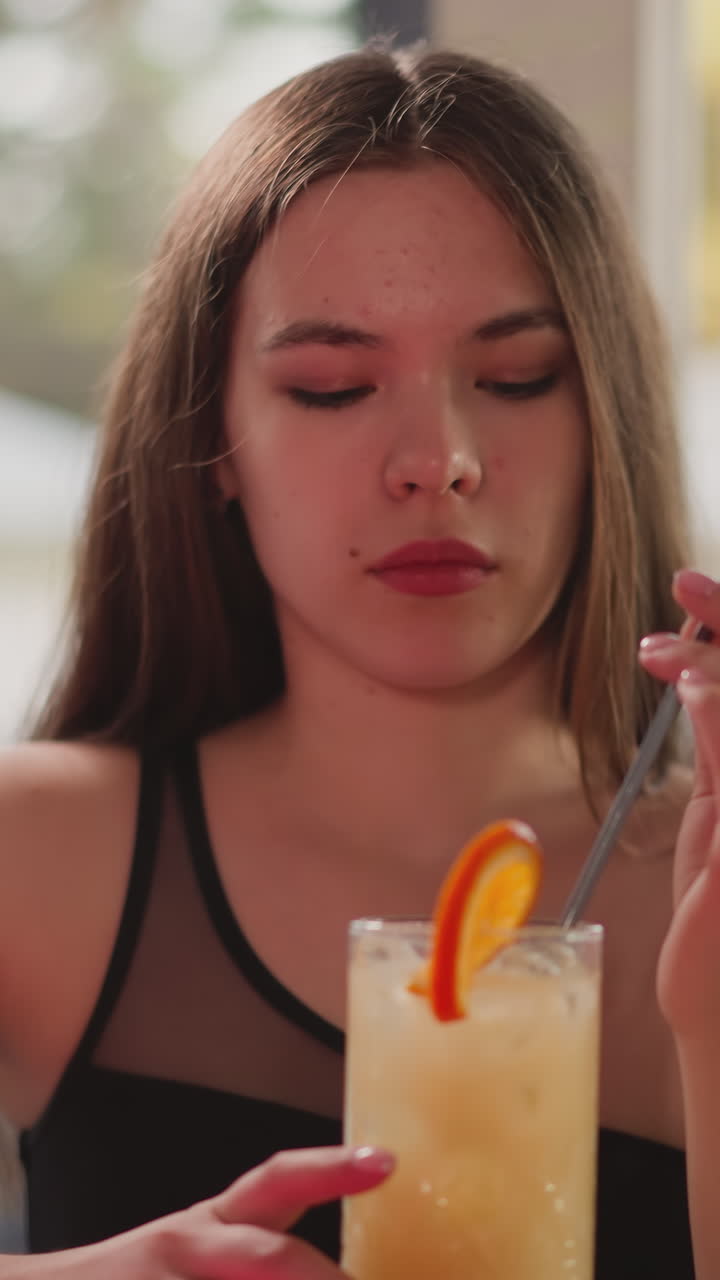 테일과 함께 지루한 여자가 바에 앉아있다. 젊은 여자가 혼자 해변 바에서 휴식을 취하는 것처럼 보입니다. 여성 손님은 오렌지 조각으로 이국적인 음료를 마시고 있습니다.