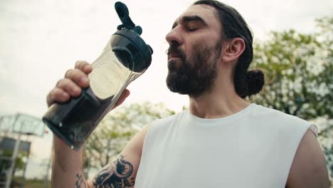 a middle-aged man with a tattoo on his arm and in a white t-shirt drinks water on the basketball court