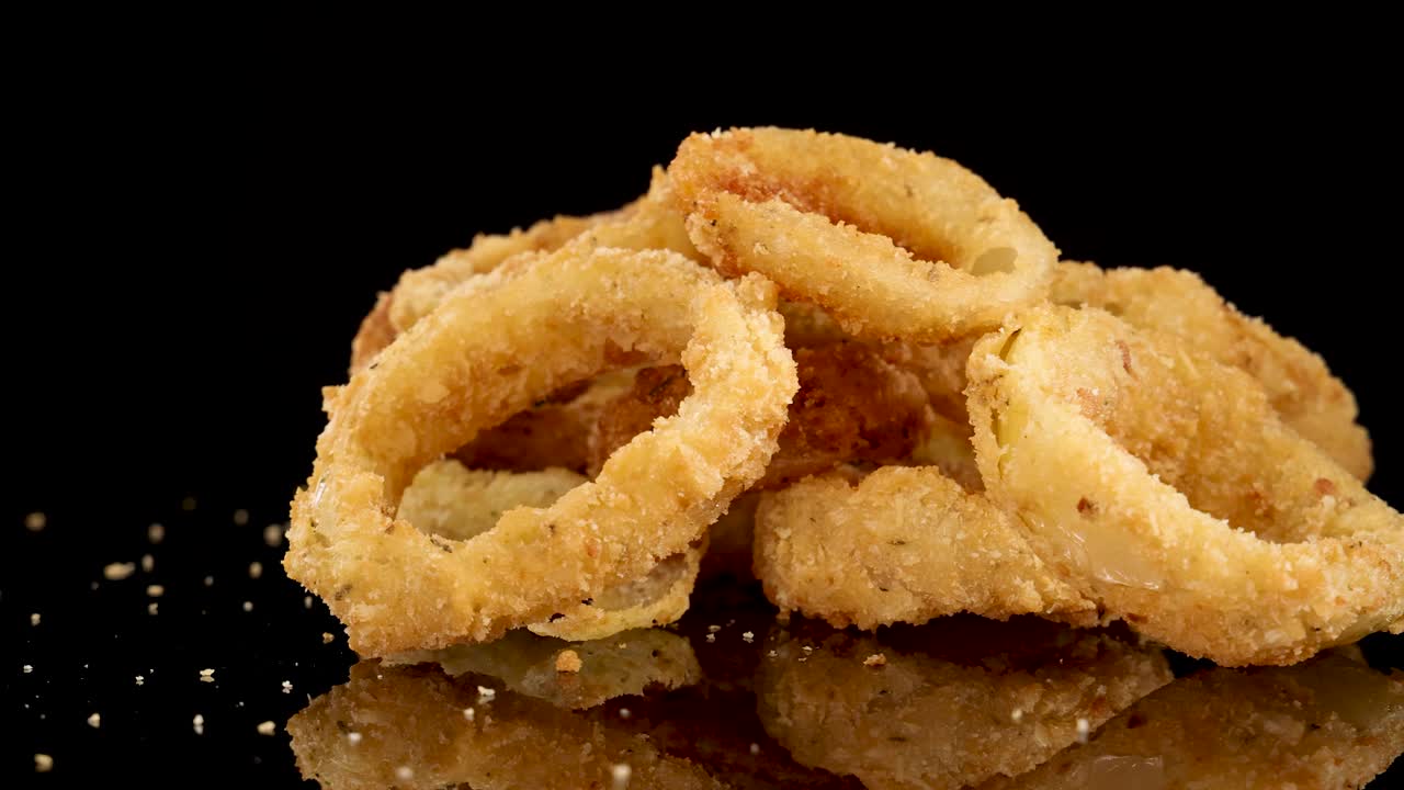 Golden fried onion rings rotate smoothly on a reflective black surface, illuminated by studio lighting, highlighting their crunchy texture and appetizing appearance