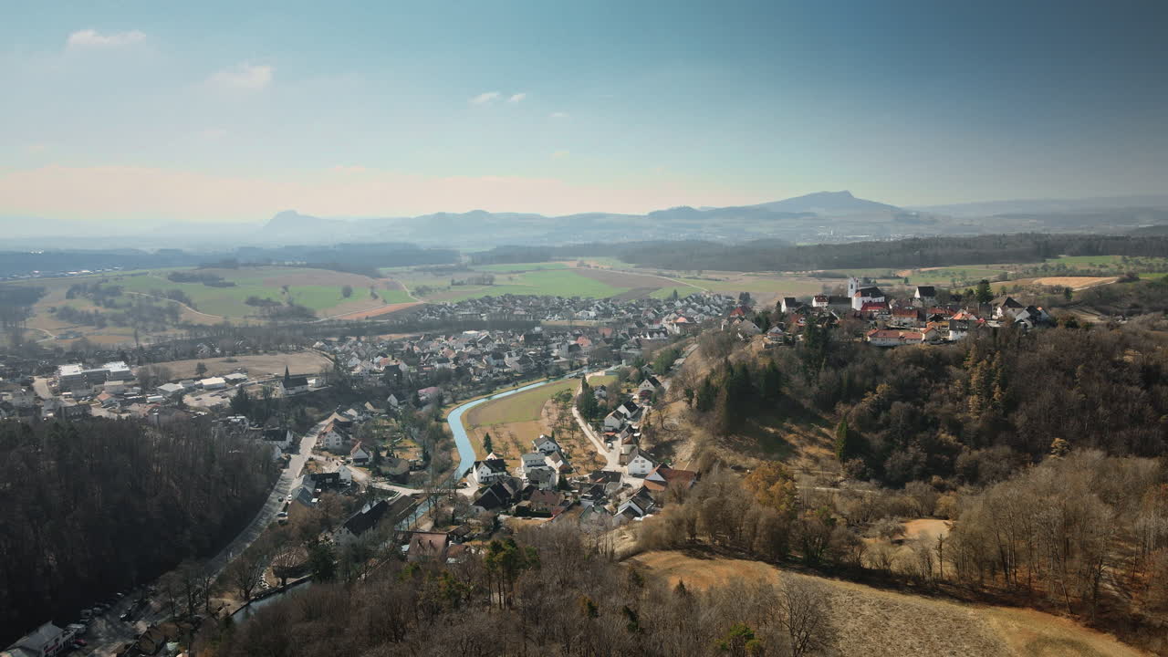 Un epico drone che si allontana all'indietro svela l'incantevole villaggio di Ulm lungo un fiume sinuoso, circondato da campi di raccolto, boschi fitti e colline lontane nella morbida luce autunnale