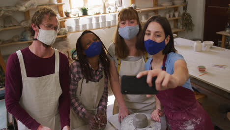 diverse potters wearing face masks and aprons taking a selfie using smartphone at pottery studio