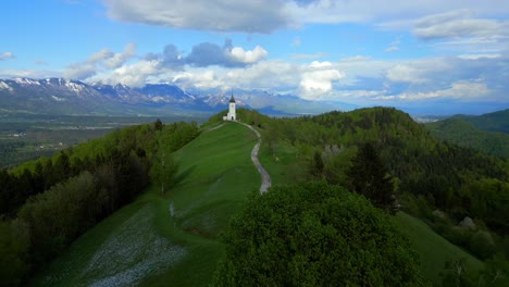 üppiger-Grüner-Hügel-Mit-Einem-Gewundenen-Pfad,-Der-Zu-Einer-Einsamen-Weißen-Kirche-Führt,-Vor-Der-Kulisse-Majestätischer-Berge-Und-Eines-Bewölkten-Himmels