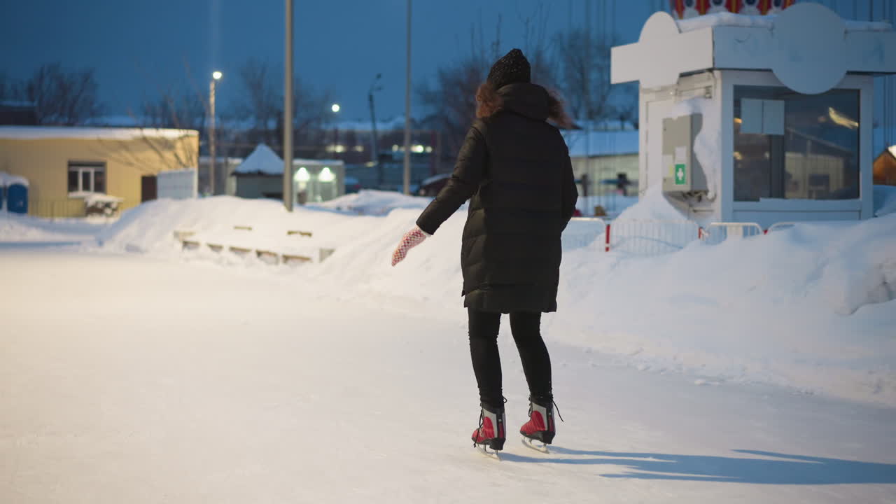 Skater gliding on outdoor ice rink wearing red skates and black coat during winter evening, surrounded by snowbanks and glowing streetlights, creating serene seasonal atmosphere filled with energy