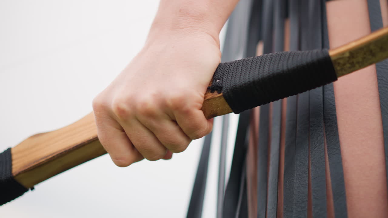 close up hand gripping leather bow, weathered wood handle wrapped in black leather, long fringe from costume cascading behind, clenched fist showing tension and readiness, knuckle texture and finger