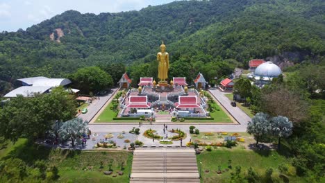 Drone video of towering golden Buddha on raised platform, surrounded by vibrant red-roofed temples amid lush forested Mae Hong Son hillside. Clear, bright lighting accentuates vibrant colors