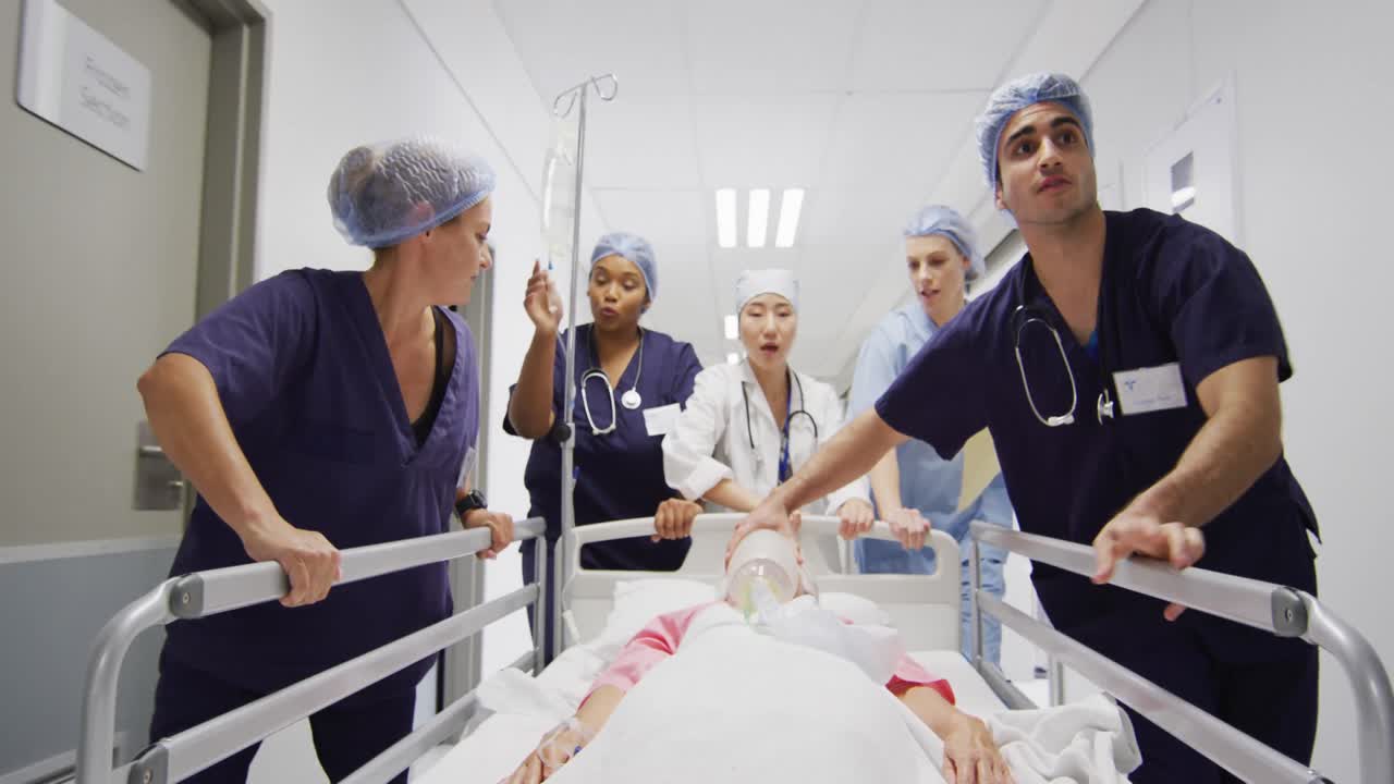 Video di un gruppo eterogeneo di operatori sanitari che spostano un paziente con ventilatore e flebo su un letto lungo un corridoio.