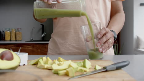 Sección-Media-De-Un-Hombre-Birracial-Mayor-Vertiendo-Batido-Saludable-En-Un-Vaso-En-La-Cocina,-Cámara-Lenta