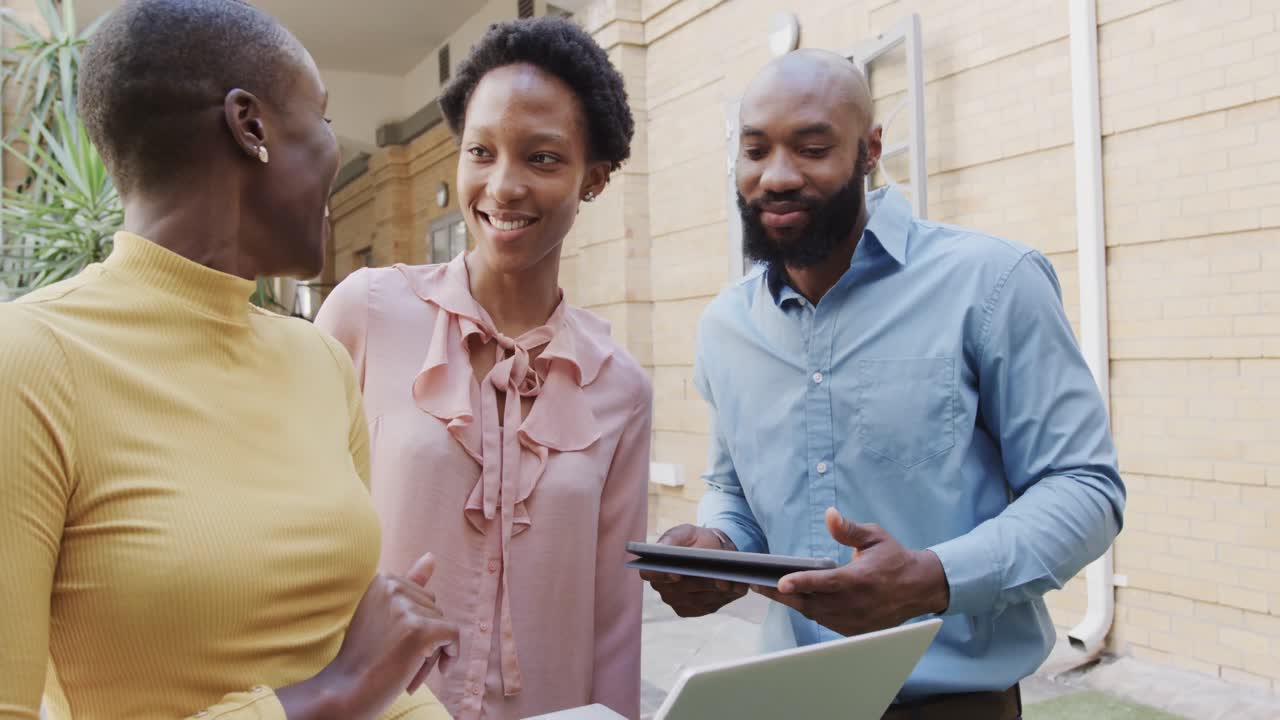 세 명의 행복한 아프리카계 미국인 남성과 여성 동료들이 밖에서 슬로우 모션으로 노트북을 사용하여 이야기하고 있습니다.