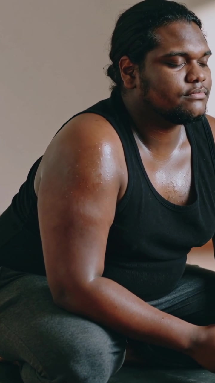 Vertical video: Breathing man in black tank and dark pants recovering after workout on mat in room