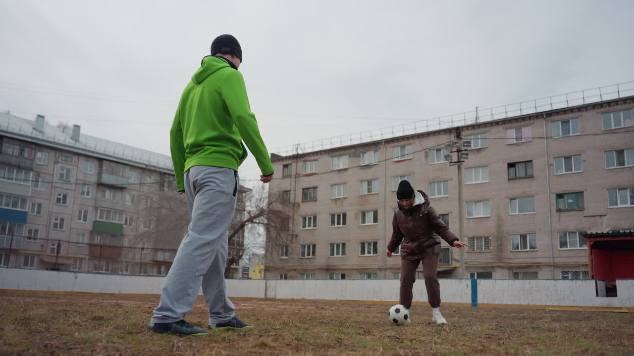 white coach and child practicing dribble and short passes on courtyard field, urban apartment blocks in background, cool light, boots and ball, patient instruction and playful repetition