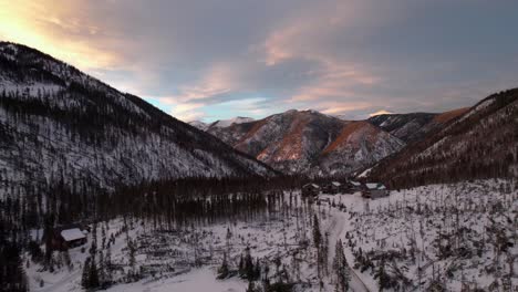 sunset drone aerial view of snowy mountains and forest with cabins