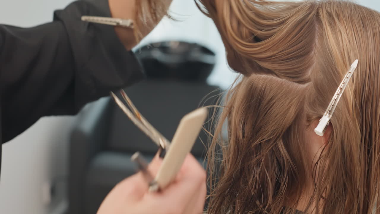 Peluquero separando el cabello castaño húmedo en secciones con un peine y clips, primer plano de las manos preparando a la clienta para un corte y tratamiento de precisión en una silla de salón moderna, luz natural suave, técnica profesional enfocada