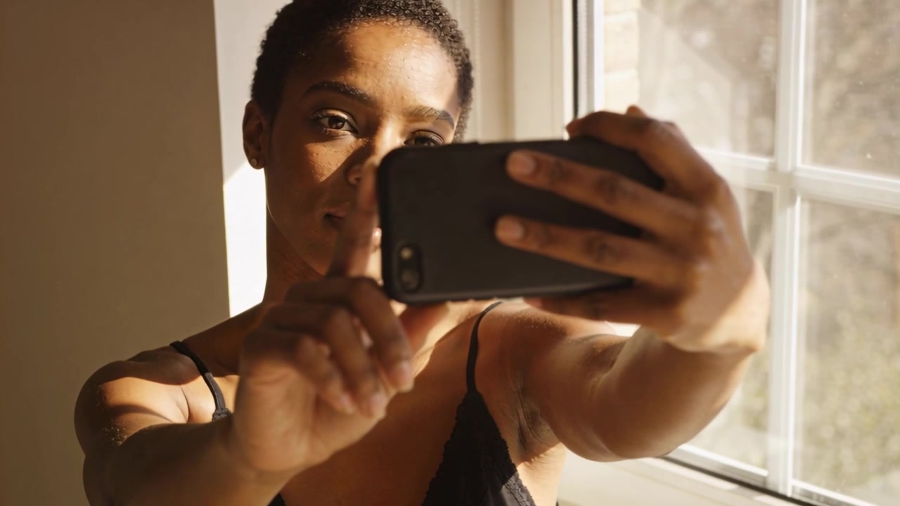 Holding blackcase phone woman in black tank angling it by paned window using light adjusting frame