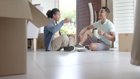 Happy-diverse-gay-male-couple-sitting-on-floor-in-new-home-toasting-with-coffees,-slow-motion