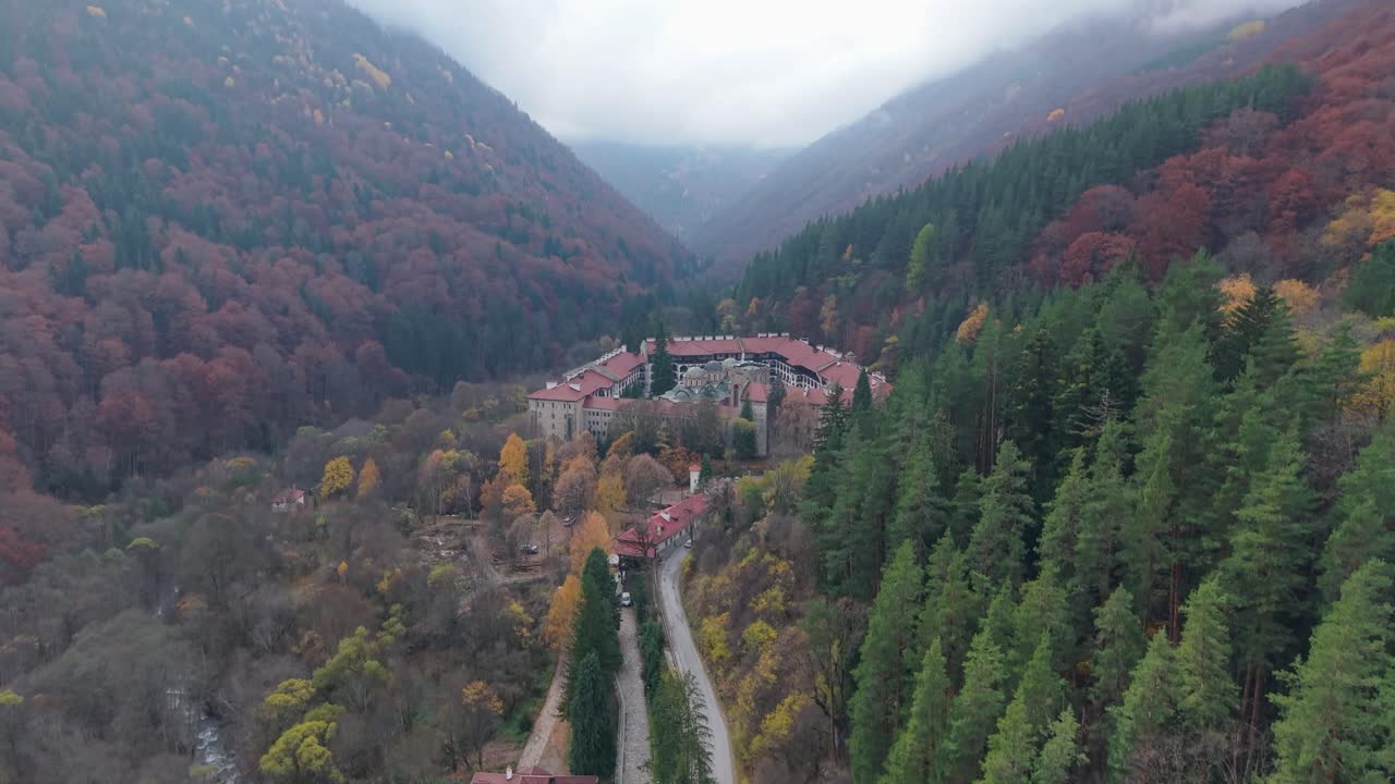 Filmato aereo con drone che segue un'auto che viaggia attraverso il paesaggio unico, rivelando gradualmente lo storico Monastero di Rila
