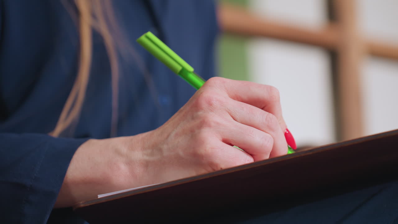 Close view of mental health professional holding green pen and notebook during client session, seated calmly with focused gesture and visible red nails, mirror in background reflects therapy space