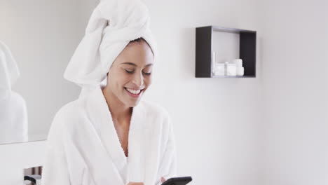 retrato de una mujer biracial feliz con una toalla en la cabeza usando un teléfono inteligente en el baño, en cámara lenta