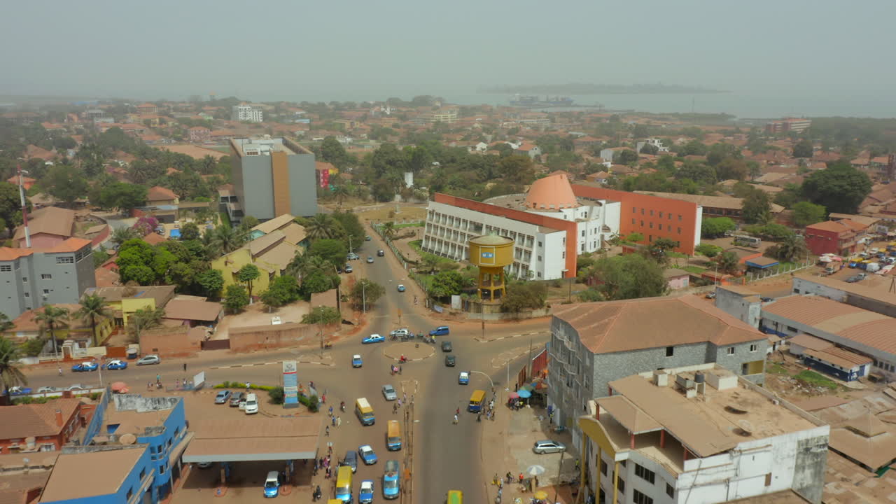 The main thoroughfare, dense low-rise architecture, and urban sprawl of a city in Guinea-Bissau, West Africa. Features traffic, market activity, and a distant water body under a hazy sky