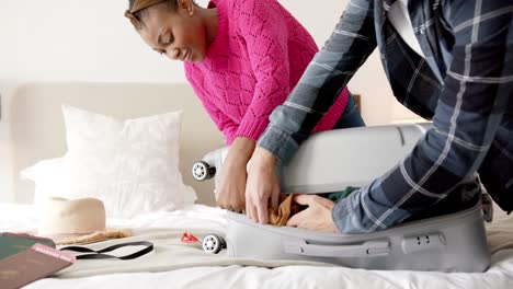 Happy-diverse-couple-packing-and-closing-suitcase-together-in-bedroom,-slow-motion