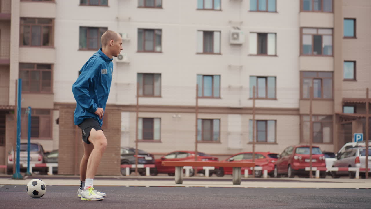 urban street soccer dribbling caucasian man in low stance with quick footwork pushing ball along wet pavement, wearing blue windbreaker and bright sneakers, apartment facades and parked cars