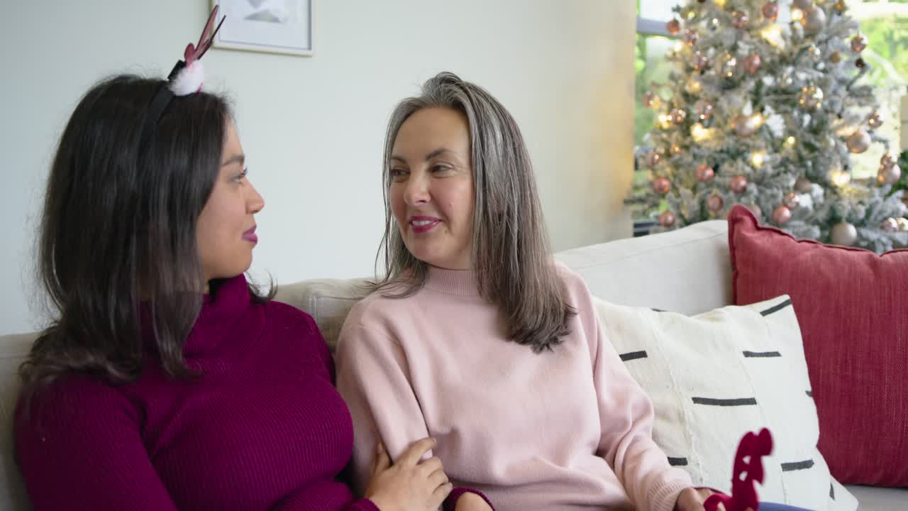 Diverse female couple settling on sofa and examining elf shoe to admire ornament by Christmas tree