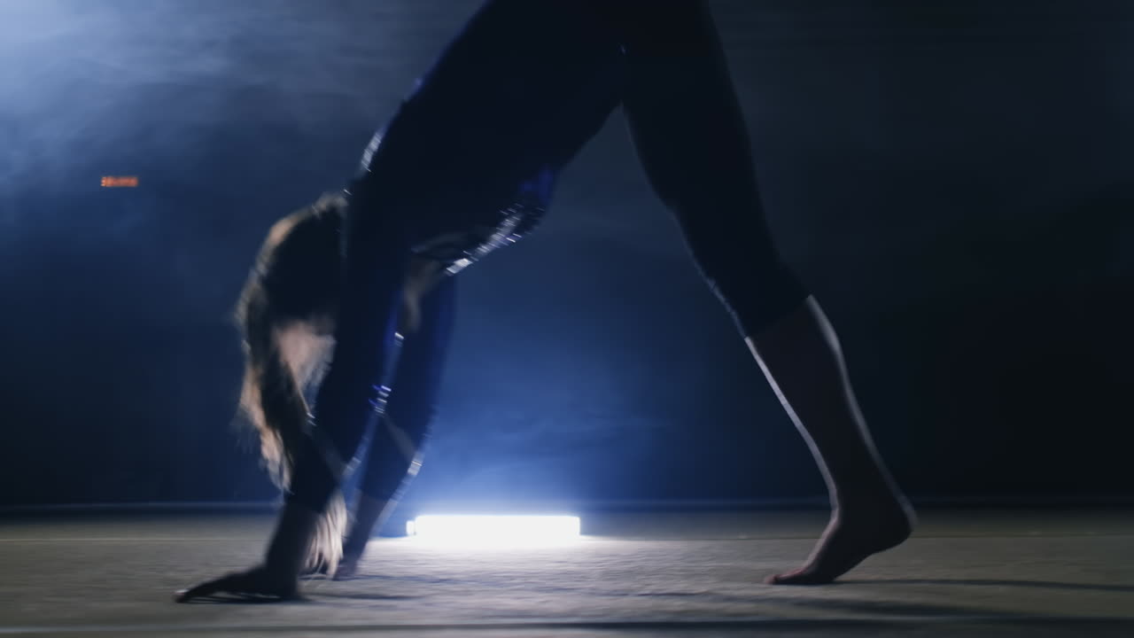 Primo piano di una ragazza ginnasta talentuosa che esegue un salto mortale sul tappeto ginnico al rallentatore, con luce sagomata nel fumo. Steadycam.